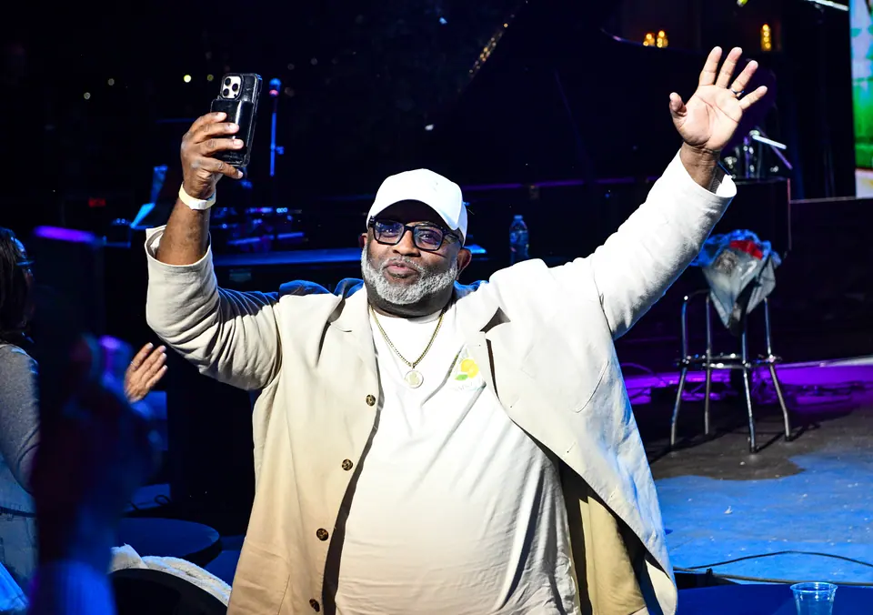 A guest in white raises his arm for a selfie with the stage behind him
