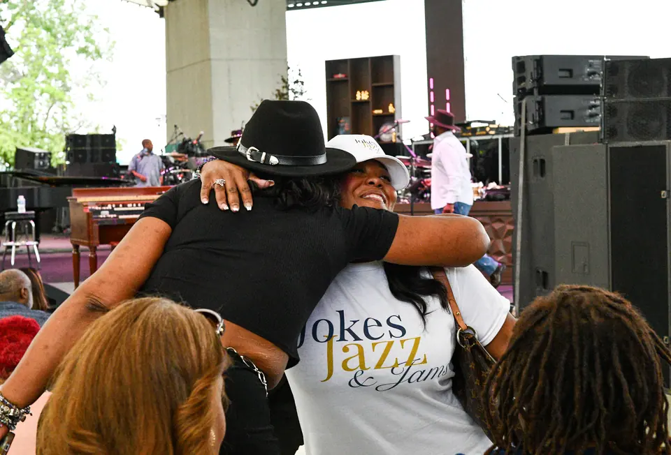 A guest in a Jokes, Jazz & Jams shirt hugs a friend near the stage
